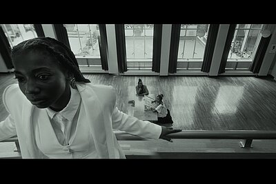 Im Vordergrund ist eine schwarze Frau in Blickrichtung zur Kamera zu sehen. Sie steht in einem Raum auf einer Galerie und stützt sich mit den Händen an der Brüstung ab. Im Hintergrund sind im Erdgeschoß 2 schwarze Frauen am Fußboden sitzend bei einem Tisch zu sehen.