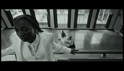 In the foreground, a black woman can be seen facing the camera. She is standing in a room on a gallery and leaning on the balustrade with her hands. In the background on the ground floor, 2 black women can be seen sitting on the floor at a table.