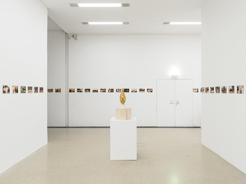 View of an exhibition room with a light-coloured floor and white walls; in the centre is a white base with a round, golden sculpture; various photos are mounted in a line on the walls