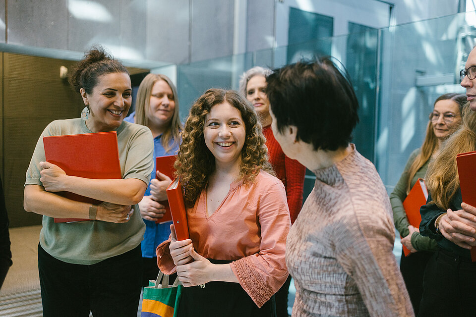 Mehrere Personen stehen mit roten Mappen in einem Eingangsbereich mit Glasflächen und Betonwänden und unterhalten sich miteinander. / Several people holding red folders stand in an entrance area with glass surfaces and concrete walls, interacting with one another.
