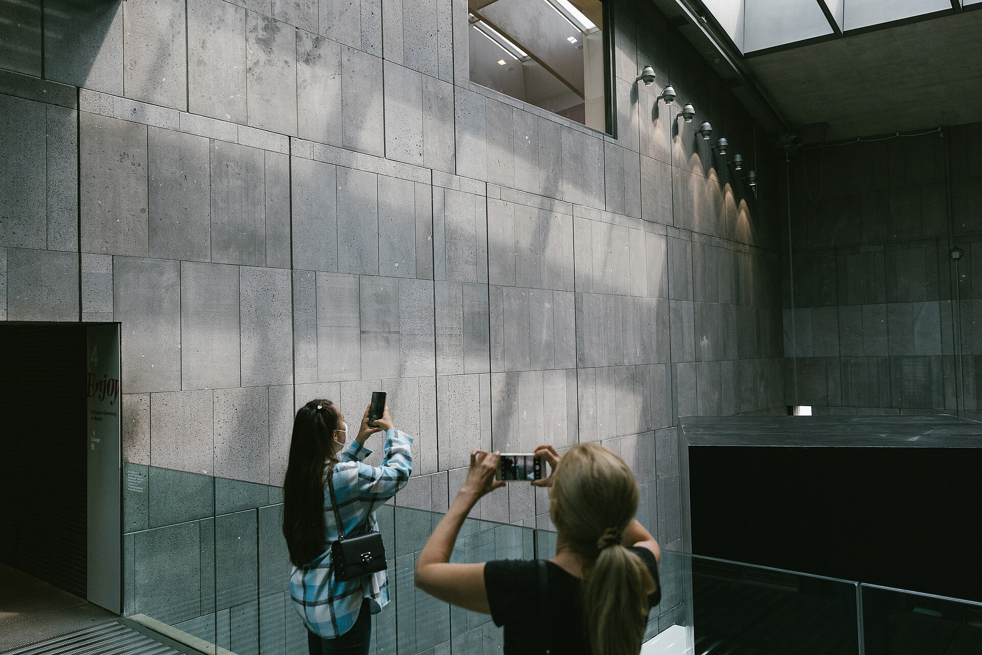 
            
                zwei Menschen stehen im mumok Treppenhaus an einer gläsernen Brüstung und fotografieren das Treppenhaus, im Hintergrund dunkelgrauer Stein an der Wand
            
        