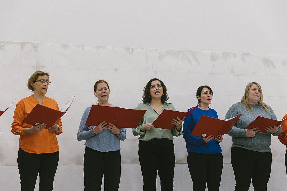 fünf Frauen in bunten Pullovern stehen in einer Reihe und halten aufgeklappte Notenbücher in den Händen, sie scheinen zu singen