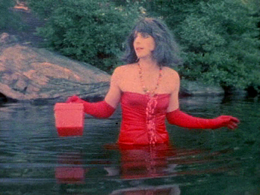 Videostill einer Person, die bis zur Taille im Wasser steht, ein rotes Kleidungsstück trägt und einen kleinen Behälter in der Hand hält, umgeben von felsiger Natur. / Video still of a person standing waist-deep in water wearing red clothing and holding a small container, surrounded by rocky natural scenery.