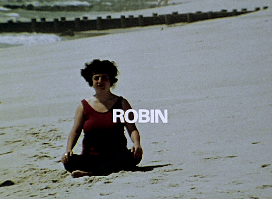 Videostill einer am Sandstrand sitzenden Person vor dem Meer; über dem Oberkörper ist der weiße Schriftzug „ROBIN“ eingeblendet. / Film still showing a person seated on a sandy beach in front of the ocean, with the white text “ROBIN” superimposed across the upper body.