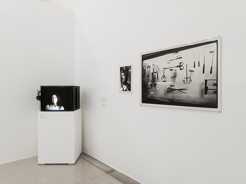 Ausstellungsansicht, links in der Ecke steht ein Fernseher auf einem weißen Sockel, rechts an der Wand hängt ein schwarz weißes Bild