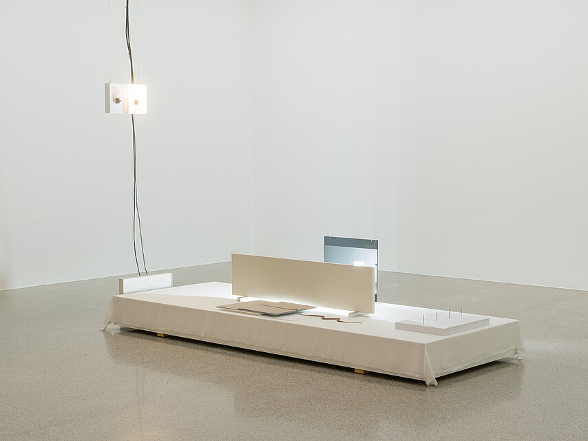 There is a light installation on a rectangular base. Hanging from the ceiling are black cables and a white box with light bulbs. 