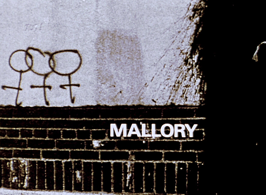 Videostill einer Mauer mit einer einfachen Zeichnung aus drei verbundenen Strichfiguren; darunter erscheint der weiße Schriftzug „MALLORY“. / Film still showing a wall with a simple drawing of three connected stick figures, with the white text “MALLORY” displayed below.