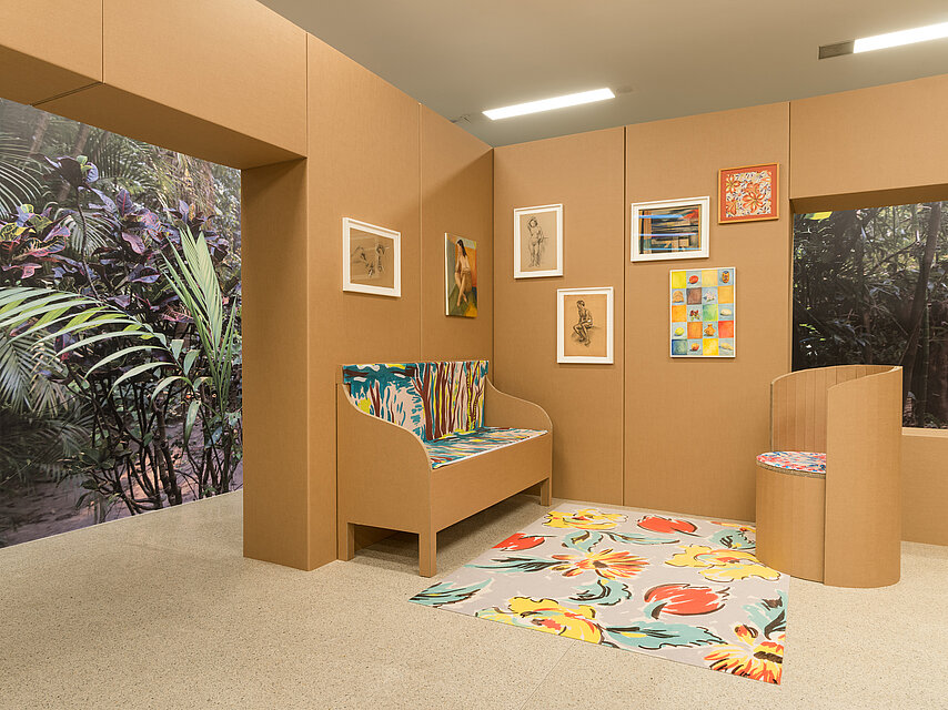 a reconstructed living room made of cardboard, on the left behind a passageway a photo of a jungle, in the middle a kind of bench, in front of it a colourful carpet, on the wall colourful works of art