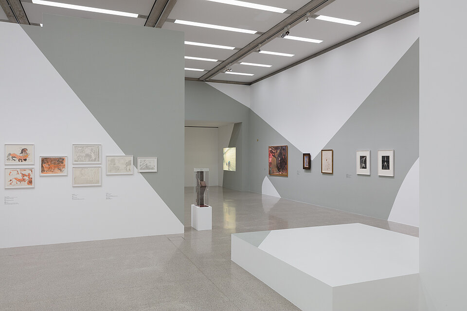 White-grey exhibition walls. Various drawings in orange and white on the left wall. On the right four more pictures. In the centre a metal sculpture on a white pedestal and an empty white pedestal next to it. Video projection in the background.