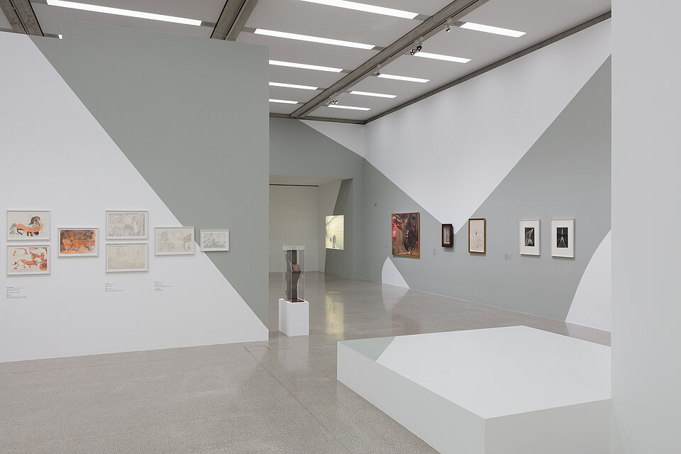 Weiß-graue Ausstellungswände. Auf der linken Wand diverse Zeichnungen in orange und weißer Farbe. Rechts vier weitere Bilder. Zentral eine Metallplasik auf einem weißen Podest sowie ein leeres weißes Podest daneben. Videoprojektion im Hintergrund.