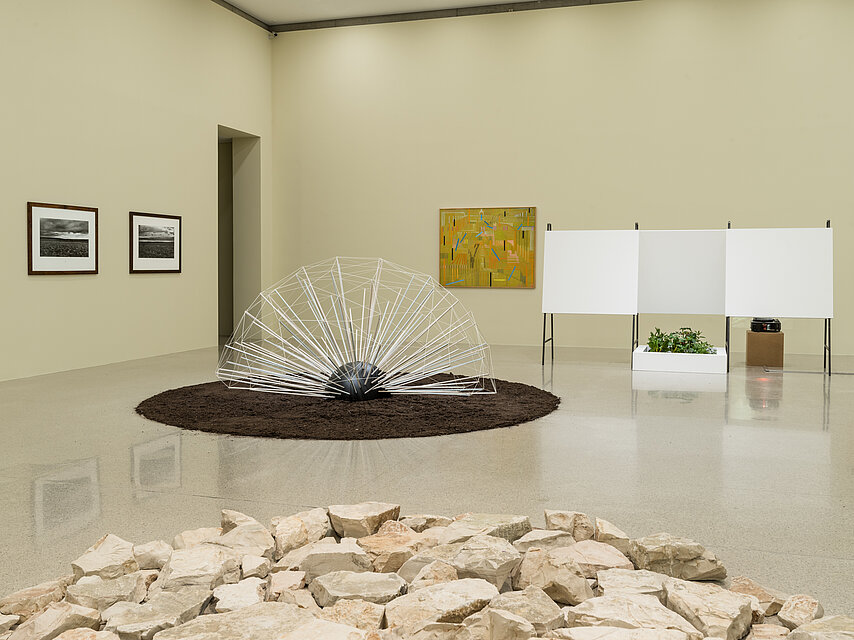 Exhibition space with light yellow walls, in the foreground light-coloured stones in a pile, in the centre an abstract object made of white, filigree material