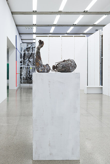 Auf einem weißen Sockel sind zwei Kunstwerke aus Metall in Form von einem Ellbogen sowie einem menschlichen Kopfplatziert.
