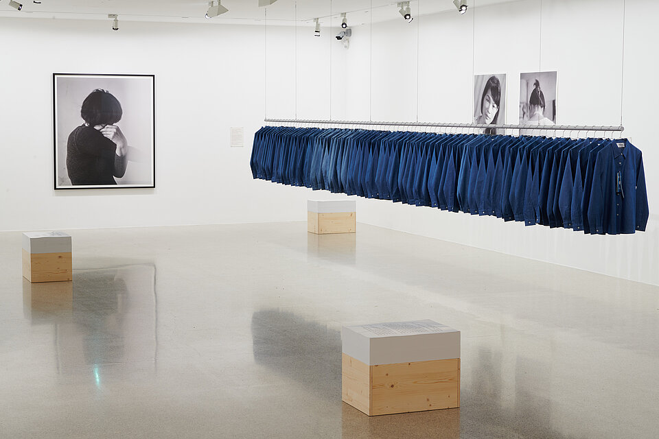 Exhibition room, on the right a long row of blue shirts hangs on a clothes rail floating in the room, on the left a portrait photo of a woman in black and white can be seen, wooden boxes stand in the room