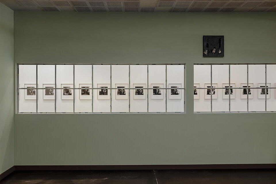 
            
                Hellgrüner Korridor mit zwei langen verglasten Wandöffnungen, durch die eine Reihe gerahmter Schwarz-Weiß-Fotografien in einem benachbarten Ausstellungsraum sichtbar ist
            
        