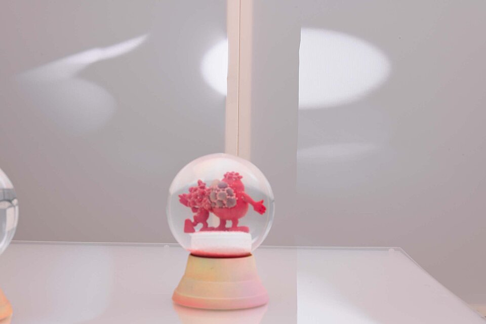 A snow globe stands on a white table. Inside the snow globe is a plastic figure in the shape of a red blood cell.