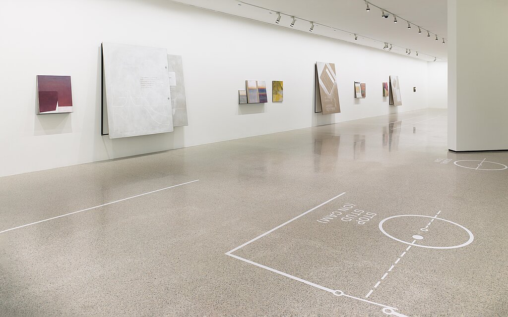 Auf der weißen Ausstellungswand hängen viele Kunstwerke in unterschiedlichen Formaten. Auf dem Fußboden befinden sich weiße Formen.