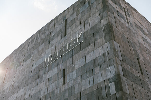 
            
                graue Fassade des mumok mit Logo "mumok", von Links strahlt die Sonne ins Bild
            
        