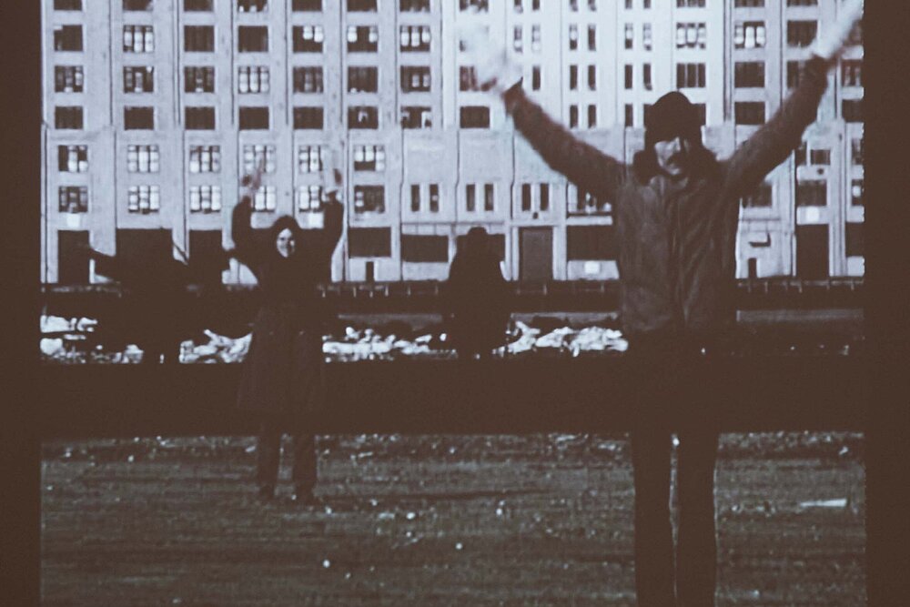 Black and white photograph of two people standing in front of a block of flats and raising their hands in the air