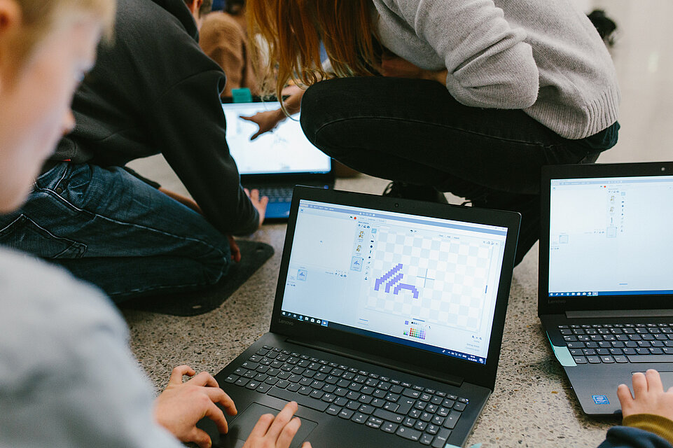 Children sit on the floor with laptops open; an animated spider can be seen on one laptop