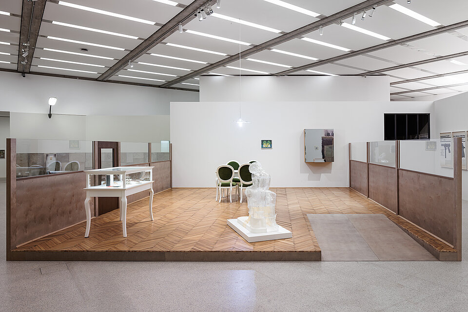 
            
                Ausstellungsinstallation auf einem erhöhten Parkettboden mit Vitrine, mehreren Stühlen, einer transparenten Skulptur und kleinformatigen Wandarbeiten
            
        