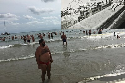 Strandszene mit vielen badenden Personen im flachen Meer unter bewölktem Himmel, überlagert von einer großflächigen Collage, die eine schwarz‑weiße Baustellenlandschaft mit eingezeichneter Figur zeigt. / Beach scene with many people in shallow water under a cloudy sky, overlaid with a large collage depicting a black-and-white construction landscape with a drawn figure.
