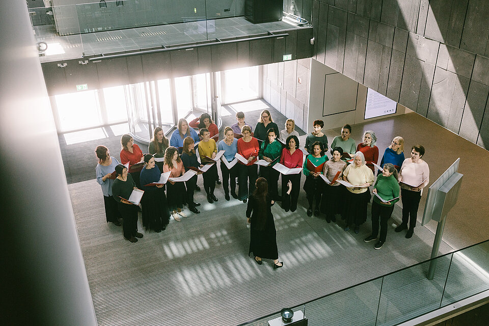 Vogelperspektive auf einen Chor, der sich mit geöffneten Mappen im Kreis um eine dirigierende Person in einer lichtdurchfluteten Museumshalle versammelt hat. / Bird’s-eye view of a choir gathered in a circle with open folders around a conductor in a light-filled museum hall.