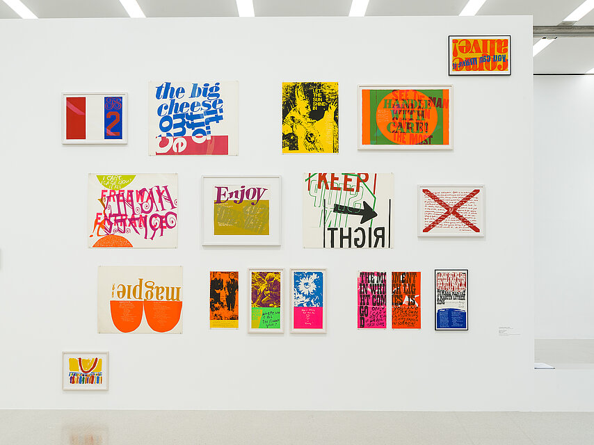 white wall with almost 15 small graphics and paintings, some of which show lettering, such as the exhibition title Enjoy
