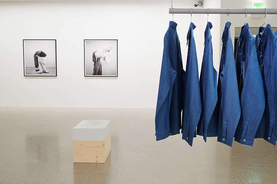 rechts blaue Hemden an einer Kleiderstange, links im Hintergrund schwarz weiße Fotografien von Menschen, vorne eine hölzerne Kiste