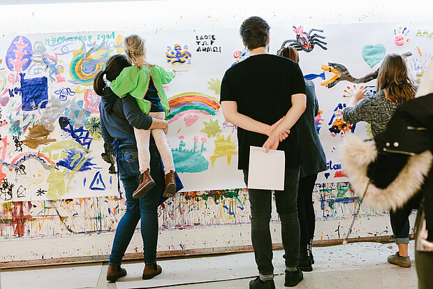 
            
                mehrere Personen und Kinder stehen vor einer weißen Wand, die voll mit bunten Zeichnungen und Farbklecksen ist
            
        