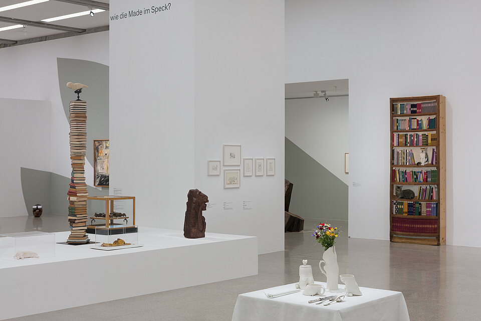 Various sculptures on a white pedestal. An installation in the form of a laid table in the foreground. 