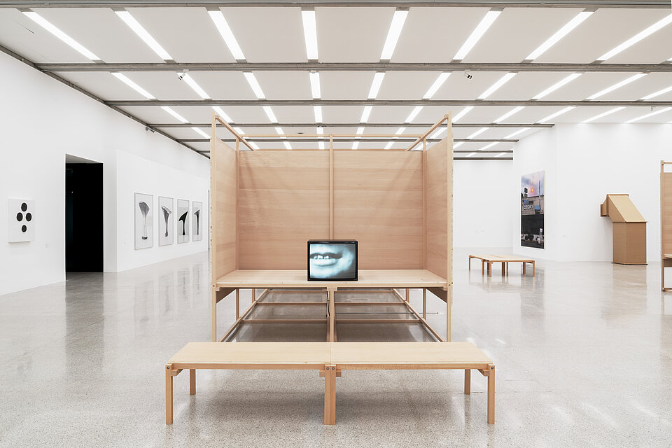 a bright exhibition room with wooden exhibition architecture, in the centre is a television showing a black and white picture