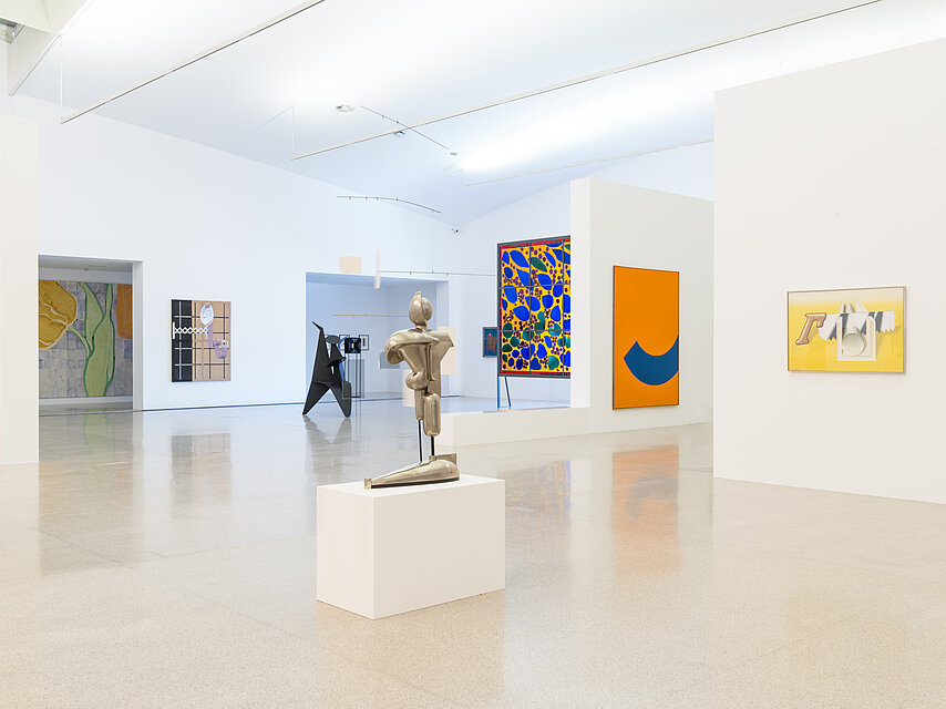 Exhibition view, a bright room with various colourful paintings, a golden sculpture on a base in the centre