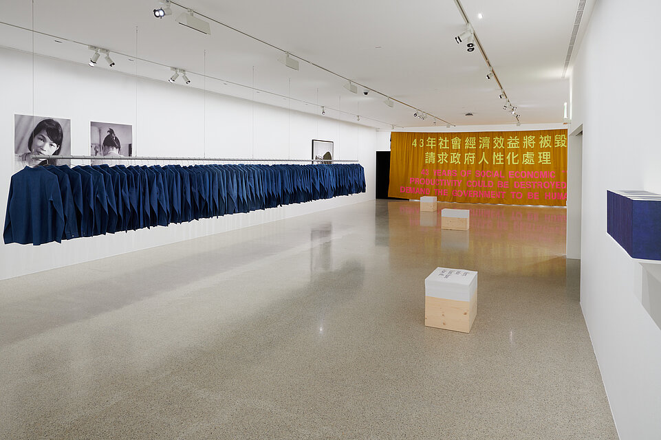 Ausstellungsraum mit einer langen Reihe blauer Hemden links an einer Stange, rechts im Hintergrund ein großer gelber Vorhang, rechts vorne eine hölzerne Kiste