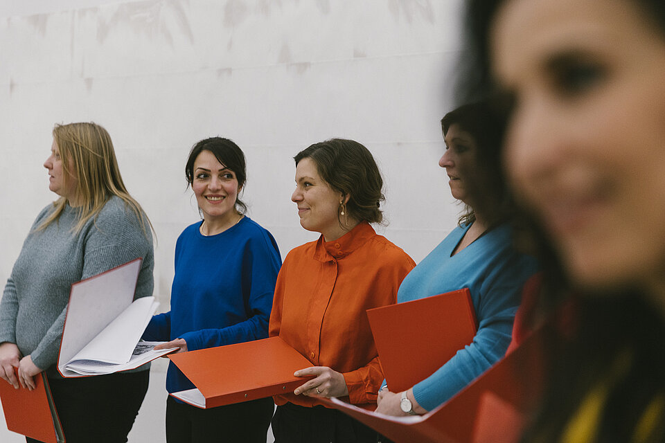 Frauen in bunten Oberteilen stehen mit Notenbüchern nebeneinander