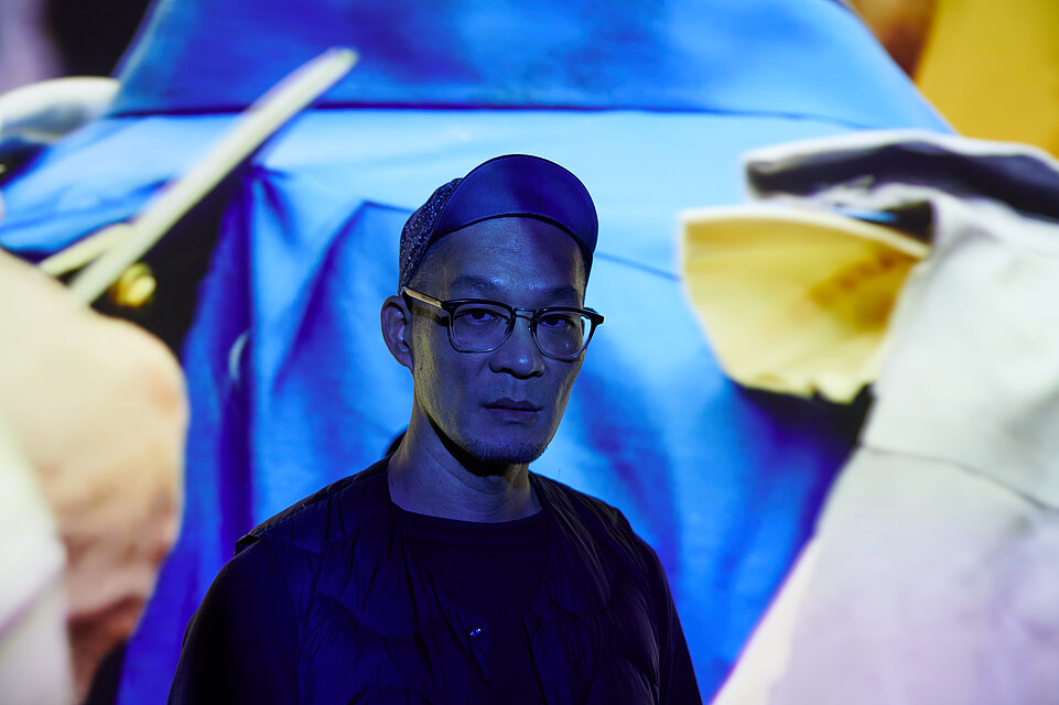 Portrait des Künstlers Huang Po-Chih, aufgenommen vor einem Projektor, der blaue Bilder auf den Künstler und den Hintergrund wirft