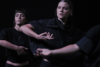 Mehrere Personen in dunkler Kleidung führen choreografierte Arm- und Handbewegungen auf einer schwach beleuchteten Bühne vor schwarzem Hintergrund aus. / Several people in dark clothing perform choreographed arm and hand movements on a dimly lit stage against a black background.