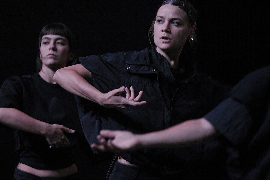 Mehrere Personen in dunkler Kleidung führen choreografierte Arm- und Handbewegungen auf einer schwach beleuchteten Bühne vor schwarzem Hintergrund aus. / Several people in dark clothing perform choreographed arm and hand movements on a dimly lit stage against a black background.