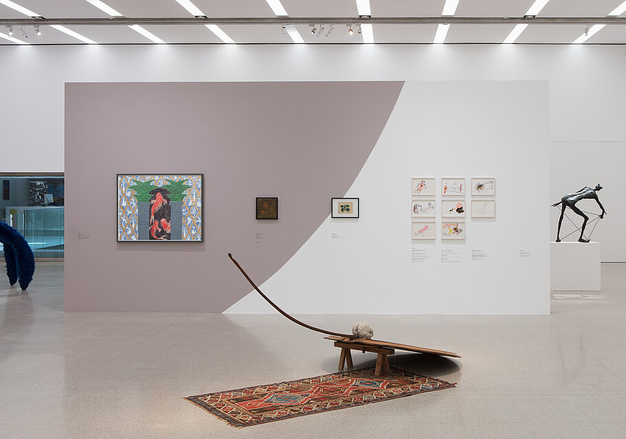 A sculpture stands on the floor in front of a white, lilac-coloured wall with paintings.