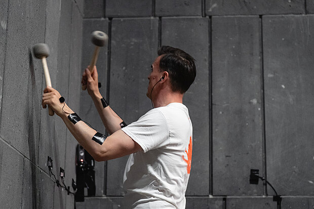 
            
                Person in einem weißen T-Shirt steht vor einer großen grauen Wand und bearbeitet die Oberfläche mit zwei Schlagwerkzeugen. Die Werkzeuge werden mit beiden Händen gegen die Wand geführt. Im Hintergrund sind industrielle Wandpaneele und Befestigungselemente sichtbar. / A person wearing a white T-shirt stands in front of a large gray wall and works on the surface using two striking tools. The tools are held with both hands and applied to the wall. Industrial wall panels and mounting elements are visible in the background.
            
        