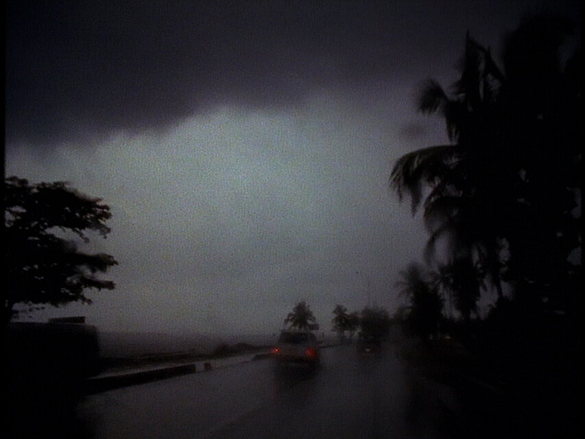 Regenverhangene Küstenstraße mit einem Auto und Palmen vor einem stürmischen Himmel. / Rain-soaked coastal road with a car and palm trees beneath a stormy sky.
