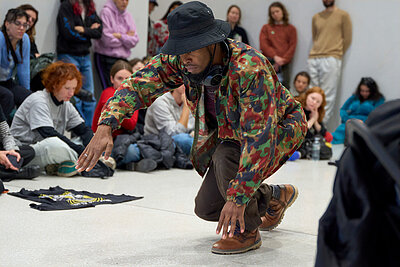 Person in gemusterter Jacke und Hut führt eine Bewegung in einem von Publikum umgebenen Innenraum aus; die Zuschauenden sitzen und stehen im Kreis um die Aktionsfläche. / Person wearing a patterned jacket and hat performing a movement in an indoor space surrounded by an audience; spectators are seated and standing in a circle around the performance area.