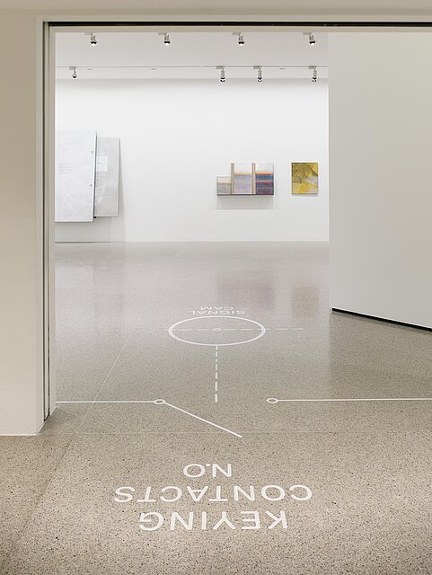 A white exhibition space with white lettering and white shapes on the floor. Three works of art hang on the exhibition wall in the background. 