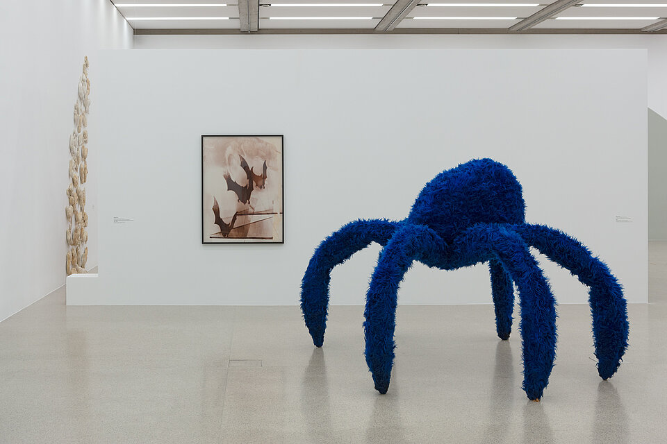 An oversized spider made of blue fur. Behind it, a work of art on the wall.