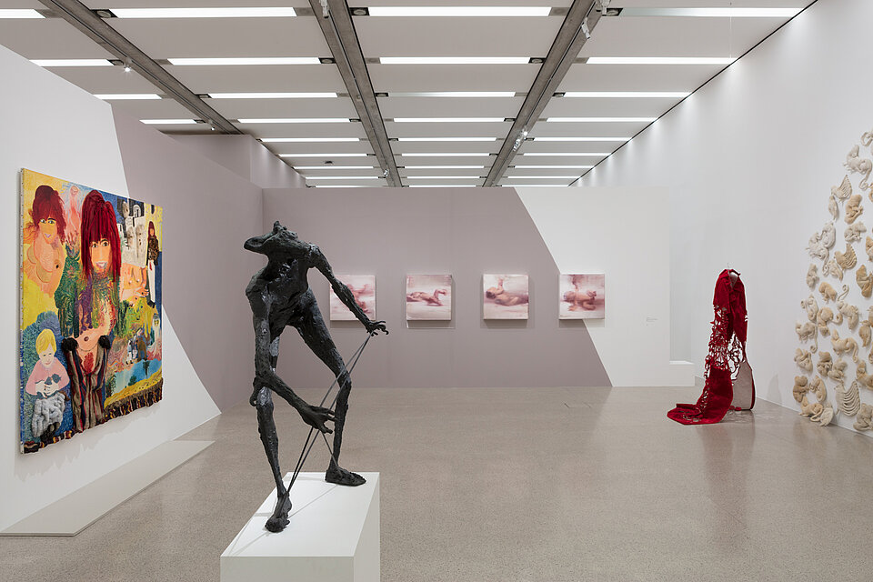 A wall installation on the right-hand side. In front of it a sculpture made of red fabric. A metal sculpture on a base in the centre. Behind it, four pictures depicting babies. A colourful picture on the left.