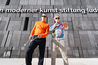 zwei Männer, der linke davon mit Drumsticks, stehen vor der mumok Fassade, oben ist zu lesen "moderner kunst stiftung lud"
