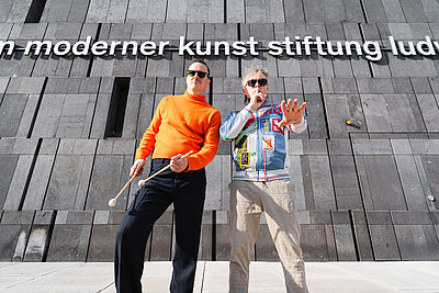 Two men, the one on the left holding drumsticks, are standing in front of the mumok façade. At the top, it reads "moderner kunst stiftung lud"