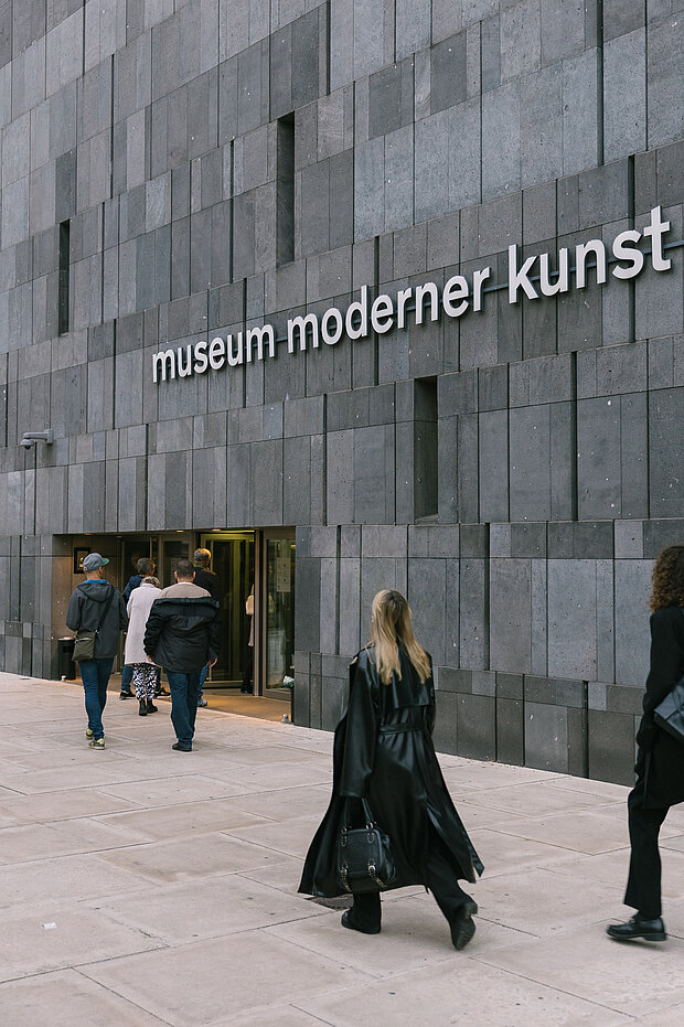
            
                Menschen vor dem mumok Eingang, die auf die Türe zu gehen, oben ist zu lesen: museum moderner kunst
            
        