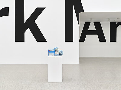 [ID: In the foreground sits a white pedestal holding a stainless steel tray filled with barrier creams, a pair of blue foam heel protectors, and individually packaged bandages. In the background, large black text partially spelling "k MAr" stretches across a white wall into an alcove leading to another section of the gallery.]