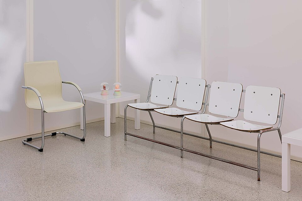  In the corner of the exhibition room, there is a small white table with two snow globes. On the left is a beige armchair with armrests. On the right is a bench with a backrest and four seats. 