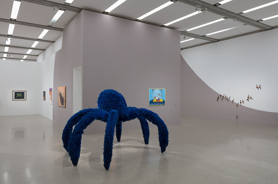  An oversized spider with six legs made of blue faux fur. Behind it, various works of art on the walls.